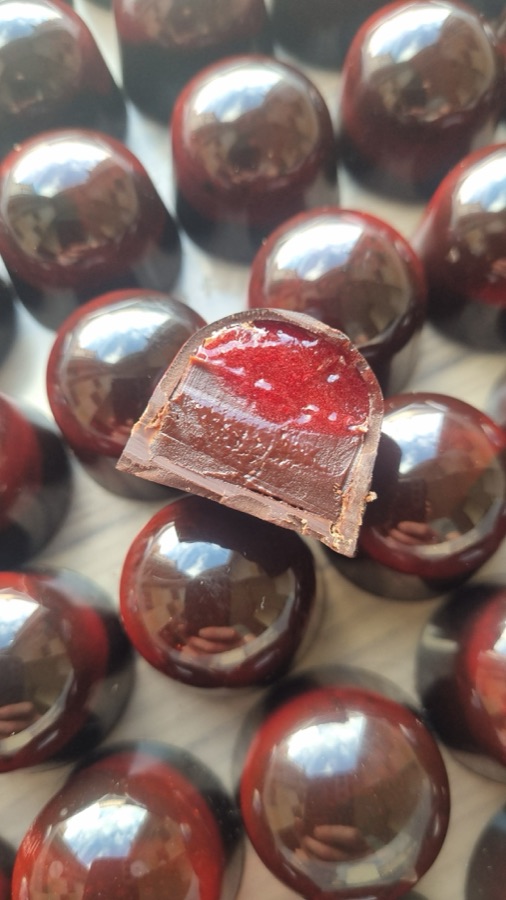 A cut bonbon showing a raspberry and dark chocolate filling, on rows of deep red bonbons