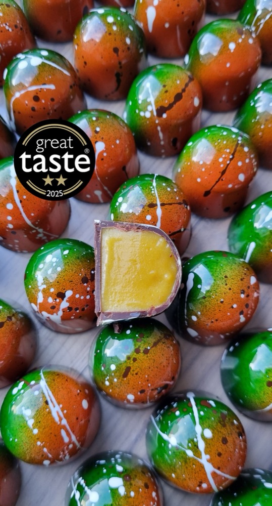 A cut chocolate bonbon showing its smooth ganache filling, resting on rows of green and orange hand-painted bonbons, with a Great Taste two-star 2025 award badge